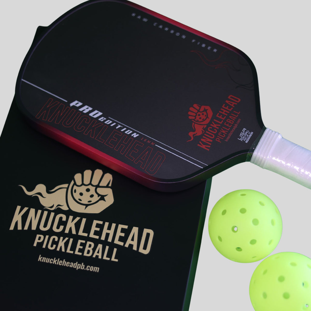 A Knucklehead pickleball paddle with a black and red face, white wrapped handle, two neon yellow pickleballs, and a black branded paddle box on a light gray background.