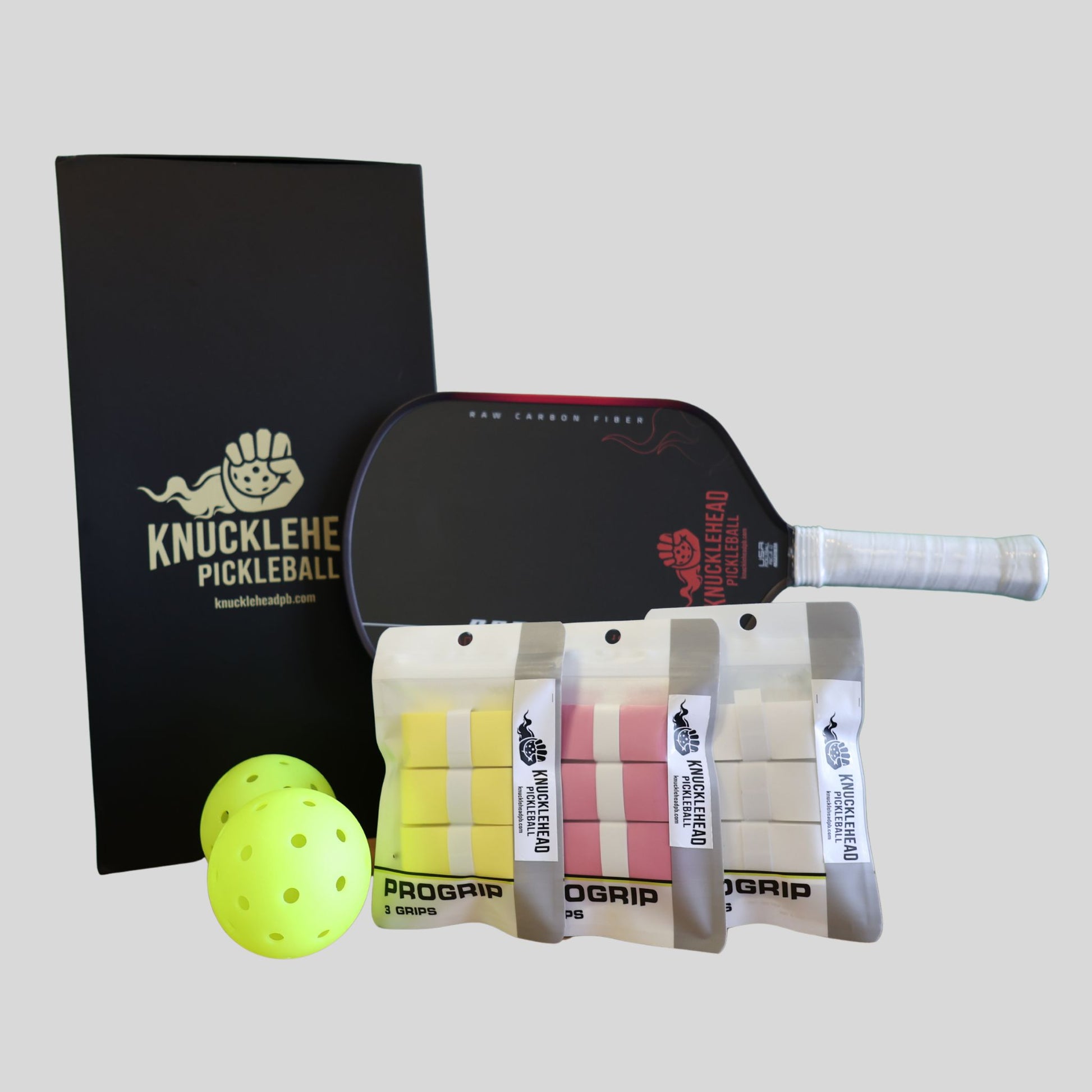 A complete pickleball kit featuring a black ProGrip pickleball paddle with a white wrapped handle, three packaged ProGrip foam balls in yellow, pink, and white, a black Knucklehead Pickleball notebook, and a bright yellow pickleball, all arranged on a light gray background.