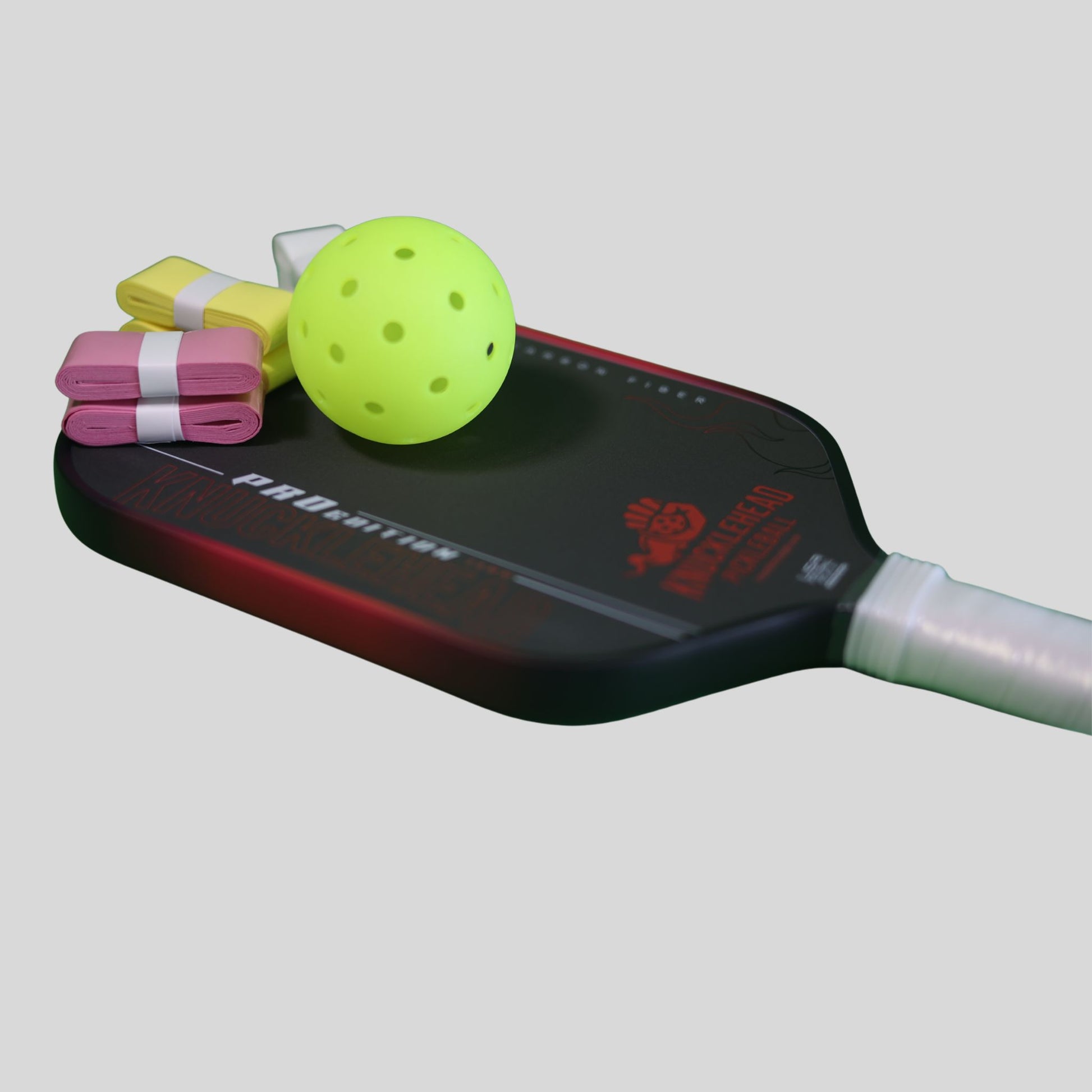 A pickleball paddle with a black and red face, shown with a neon yellow pickleball ball and folded colored paddles on top, against a solid gray background.