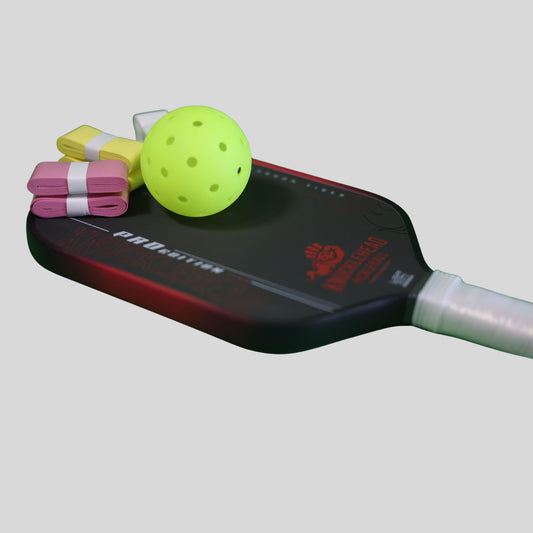 A pickleball paddle with a black and red face, shown with a neon yellow pickleball ball and folded colored paddles on top, against a solid gray background.